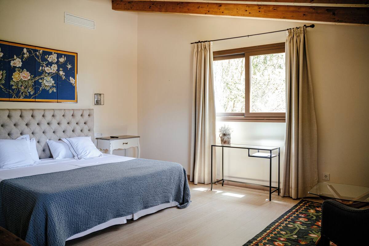 Un dormitorio con una cama grande, una ventana con cortinas, una mesita pequeña y un cuadro decorativo en la pared.