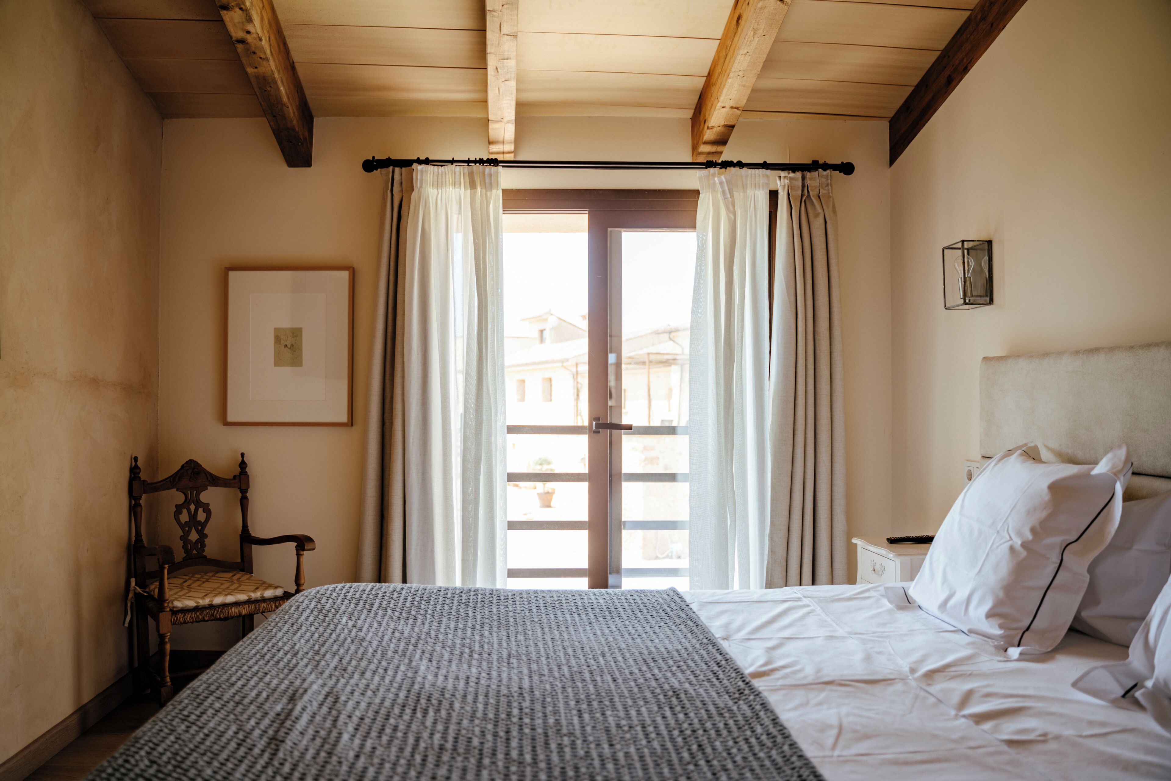 Un dormitorio con cama, una silla, una mesita de noche, un marco de foto y una ventana con cortinas.