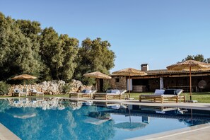 Une piscine avec des chaises longues et des parasols, un mur en pierre et un bâtiment doté d’un toit en tuiles.