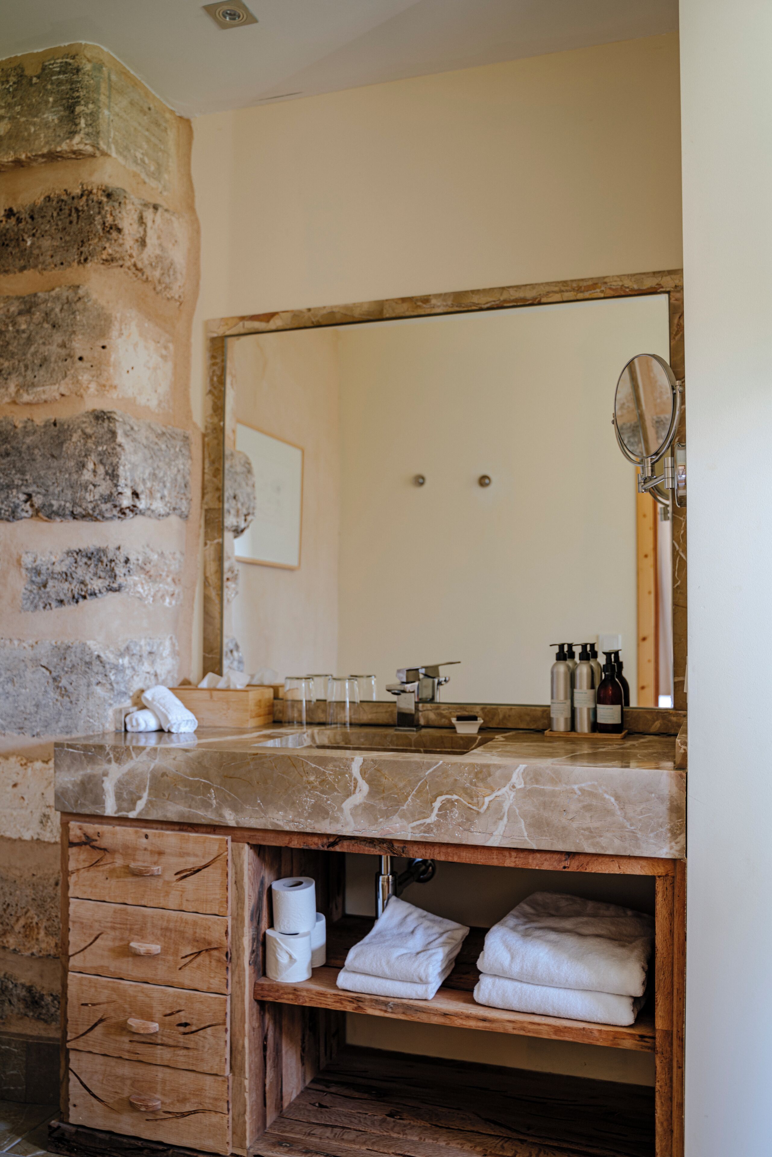 A bathroom with a stone wall, wooden vanity, marble sink, and a large mirror.