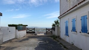 A narrow alleyway leading to a beach with a gate, a white building with blue shutters, and a clear view of the sea. - La Luciole – 3★ bright & comfortable apartment by the sea (ronce les bain)