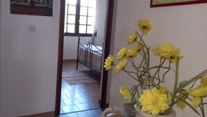 A room with a bed, a window with curtains, a painting on the wall, and a vase with yellow flowers on a table. - Charming house near the sea South Corsica (Sollacaro)