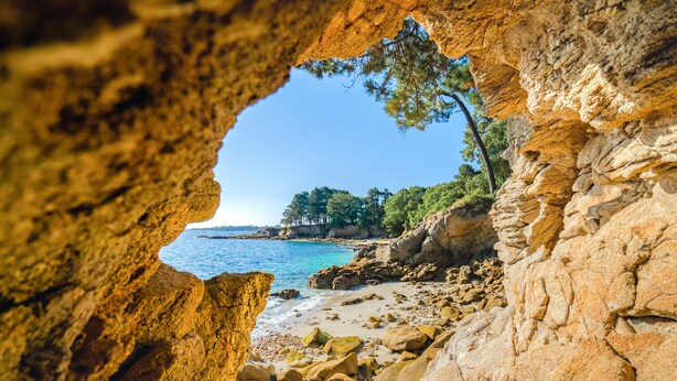 Une vue sur la côte, encadrée par une arche naturelle formée par des rochers, avec une plage et des arbres en arrière-plan.
