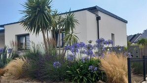 Une maison moderne à deux étages avec un garage, entourée d’une grande variété de plantes et de fleurs.