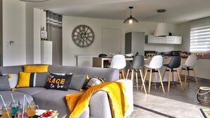 Un salon moderne avec un canapé gris, des coussins jaunes et un coin repas comprenant des chaises blanches et une table en bois.