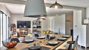Un espace de salle à manger moderne avec une longue table en bois, dressée pour six personnes, des luminaires suspendus au plafond et une bibliothèque en arrière-plan.