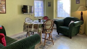 Ein Wohnzimmer mit einem grünen Sofa, einem Esstisch mit Stühlen, einem an der Wand montierten Fernseher und einem Fenster mit Blick auf Gebäude.