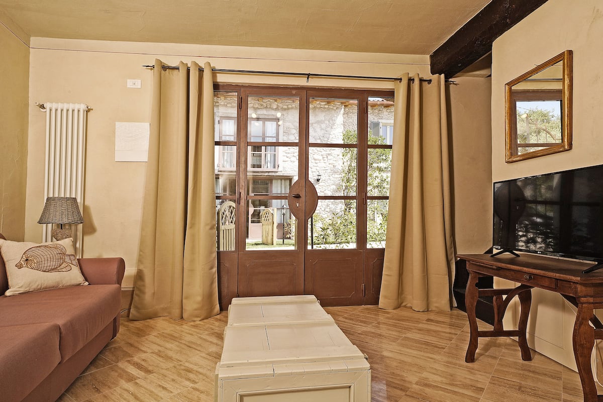 A room with a wooden floor, a brown sofa, a white ottoman, a wooden table, and a window with curtains.