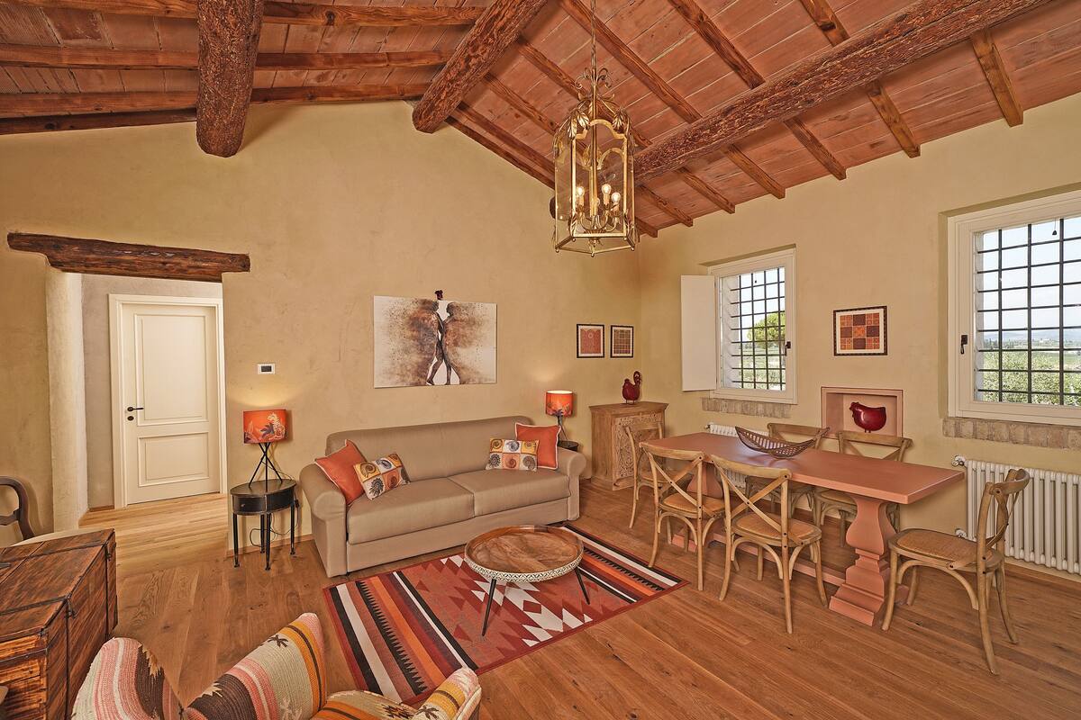 A cozy living room with a wooden ceiling, a sofa, a dining table with chairs, and a large window.
