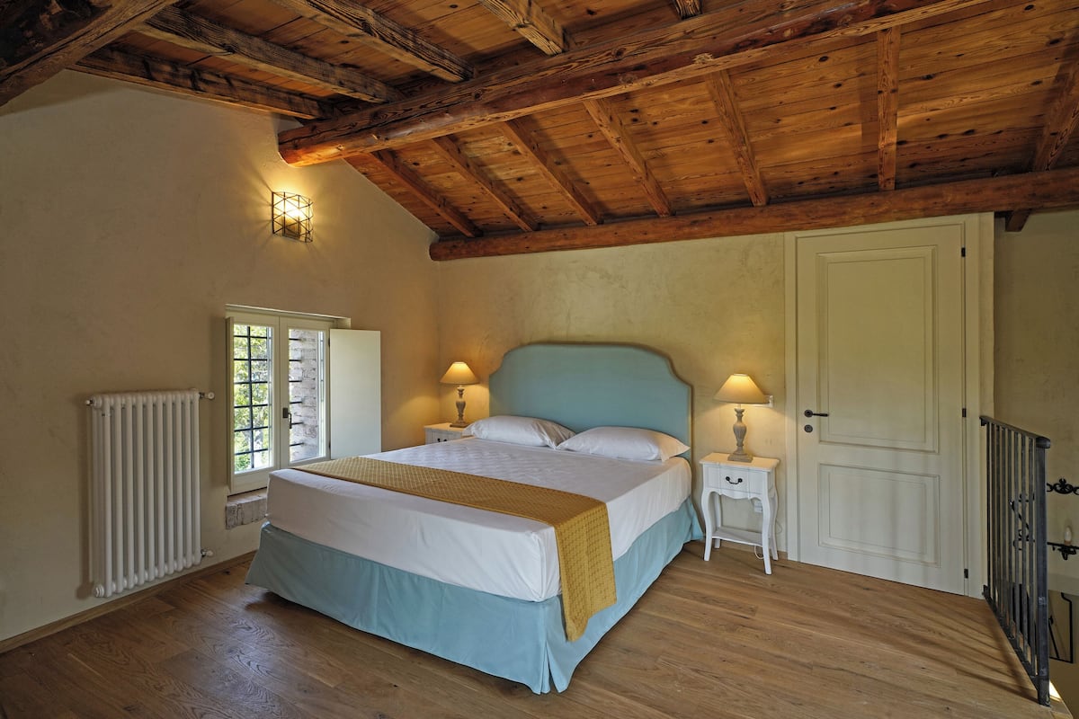 A bedroom with a wooden ceiling, a bed with white bedding, a nightstand with a lamp, a radiator, and a door.