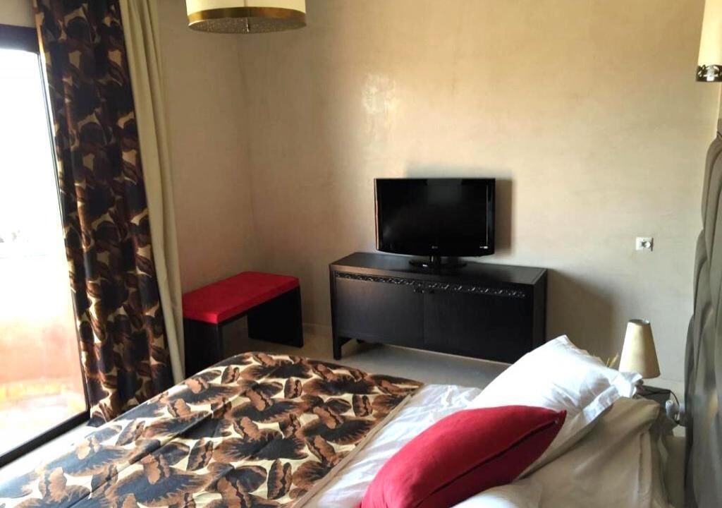 A hotel room with a bed, a television on a dresser, a red bench, and a window with curtains.