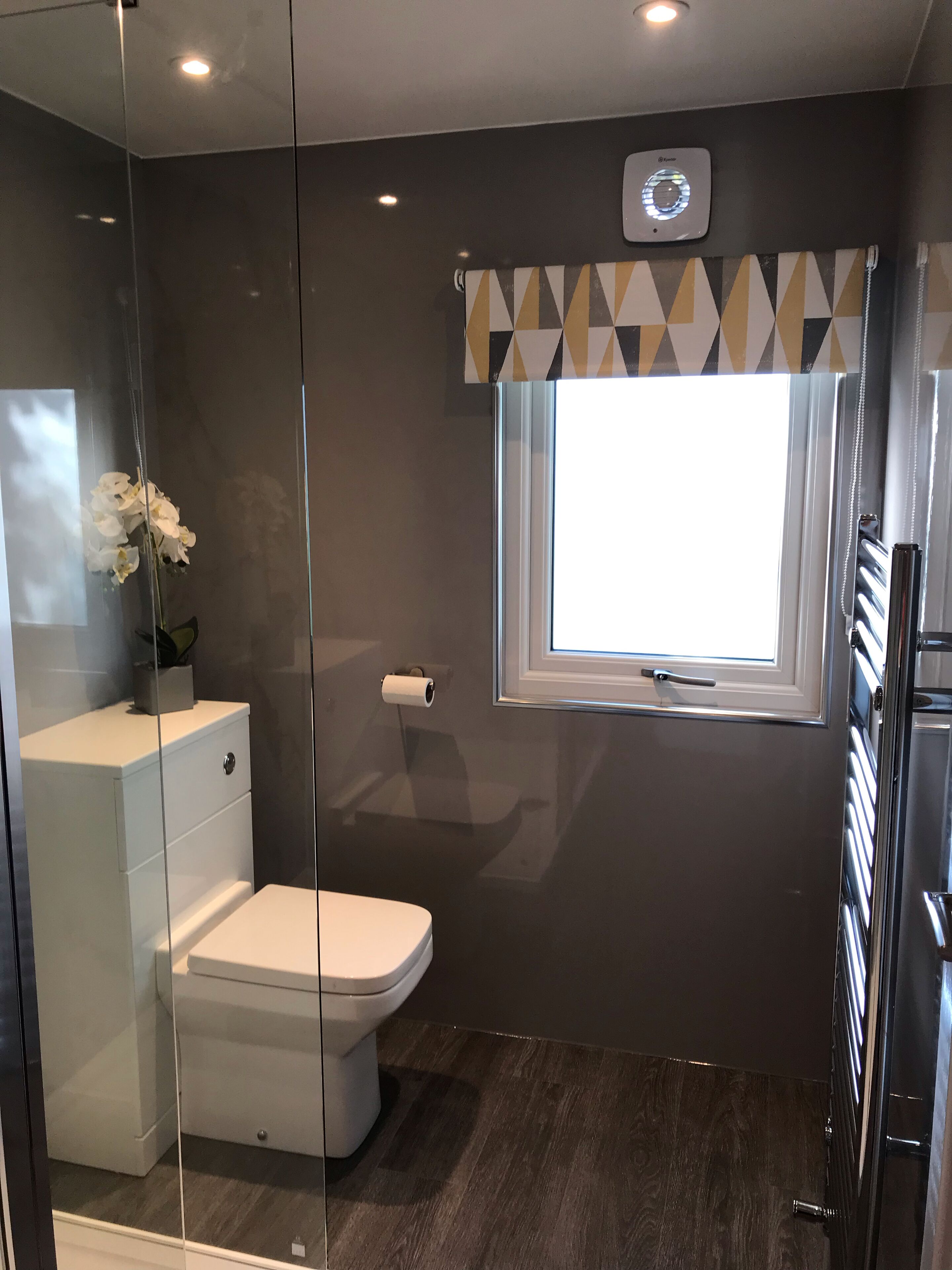 Un baño moderno con inodoro, cabina de ducha con mampara de cristal, ventana con cortina geométrica y dispensador de papel higiénico en la pared.