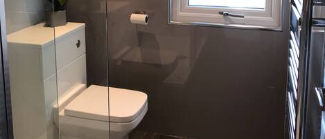 Un baño moderno con inodoro, cabina de ducha con mampara de cristal, ventana con cortina geométrica y dispensador de papel higiénico en la pared.