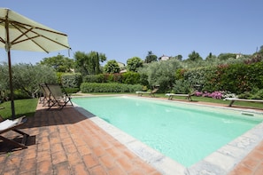 A rectangular swimming pool with a tiled edge, surrounded by a brick patio. There are lounge chairs and a large umbrella on the patio. - Maestà del Sasso (Cortona)