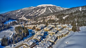 Un complejo turístico en la montaña cubierto de nieve, con edificios de estilo chalet dispuestos en filas, una pista de esquí y montañas boscosas en el fondo.