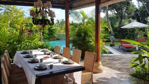 Un espace de restauration en plein air avec une table dressée pour six personnes, entouré de chaises en osier, sous une pergola en bois agrémentée de lampes suspendues. Une piscine et une végétation luxuriante sont visibles au fond.