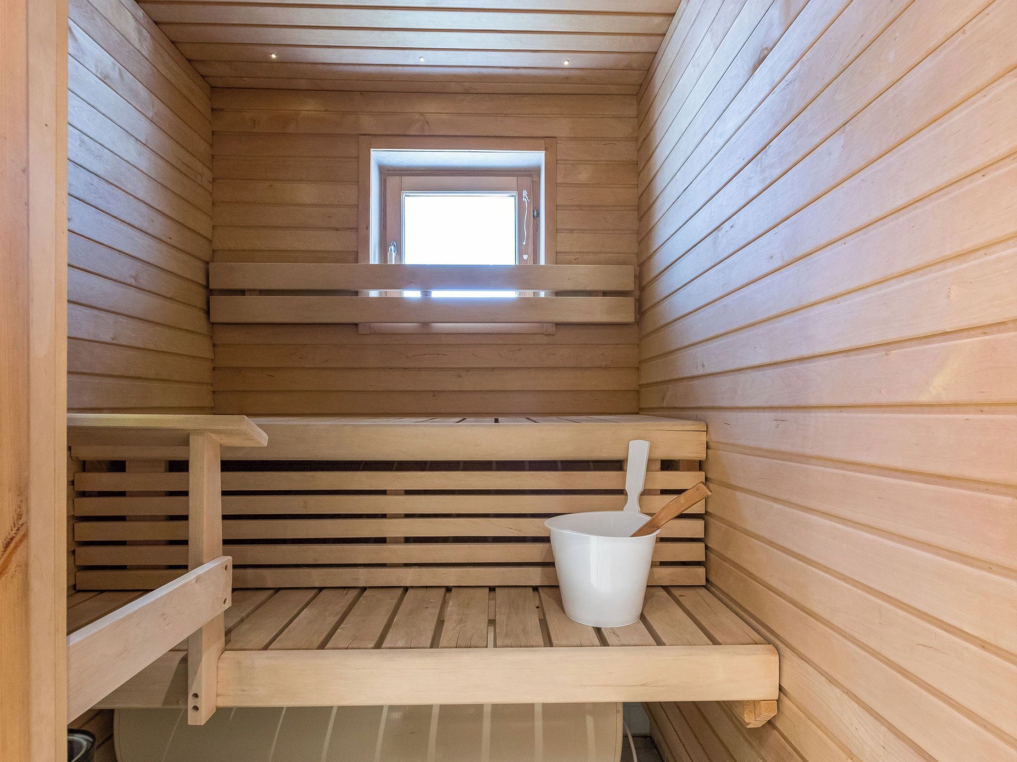 A sauna with wooden benches, a small window, and a white bucket.