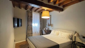 A bedroom with a wooden ceiling, a bed with white bedding, a bedside table with a lamp, and a TV on the wall. - Le camere di Bsuites (Parma)