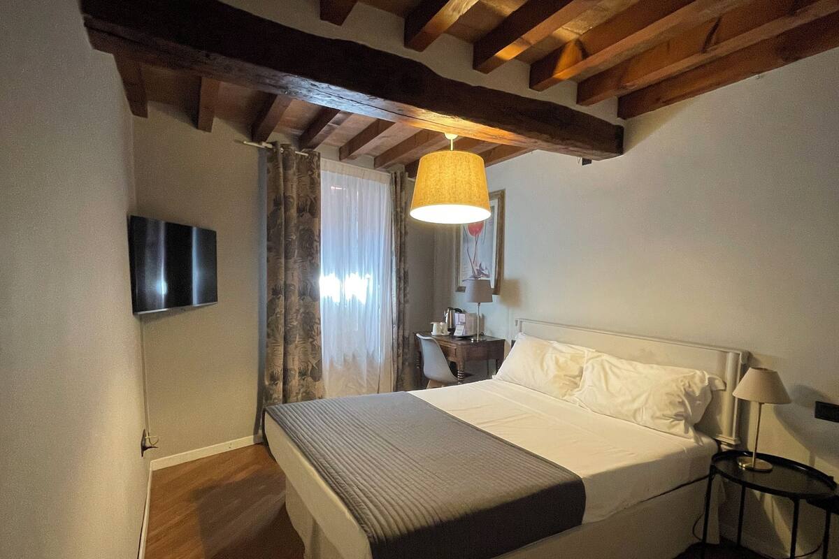 A bedroom with a wooden ceiling, a bed with white bedding, a bedside table with a lamp, and a TV on the wall.