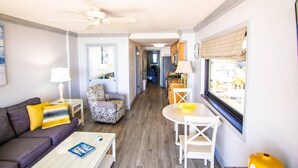 A compact living space with a sofa, a dining table, a patterned armchair, and a ceiling fan. A kitchen area is visible in the background. - Beautiful End Unit Oceanfront in the heart of Myrtle Beach indoor and outdoor pools 203 (Myrtle Beach)