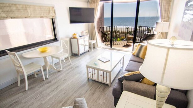 A hotel room with a balcony, a TV, a dining table, and a sofa. - Beautiful End Unit Oceanfront in the heart of Myrtle Beach indoor and outdoor pools 203 (Myrtle Beach)