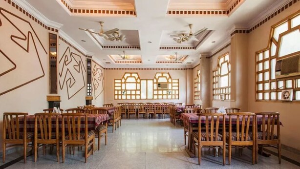 A spacious dining hall with high ceilings, chandeliers, and a central open space surrounded by wooden tables and chairs. - Hotel Saurab (Dehradun)