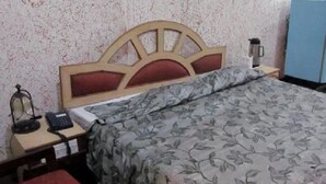 A bed with a floral bedspread, a wooden headboard, a bedside table with a lamp, a phone, and a coffee maker. - Hotel Saurab (Dehradun)