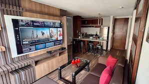 A modern living room with a flat-screen TV displaying a streaming service interface, a kitchen area with bar stools, and a cozy seating area with a sofa and a coffee table. - LIMA FLAT 2 1D BETWEEN AIRPORT AND MIRAFLORES (San Miguel)