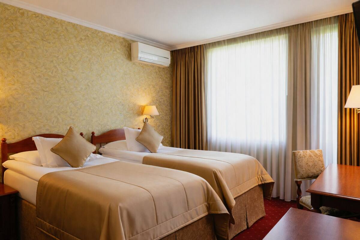 A hotel room with two beds, a desk, a chair, a lamp, and an air conditioning unit.
