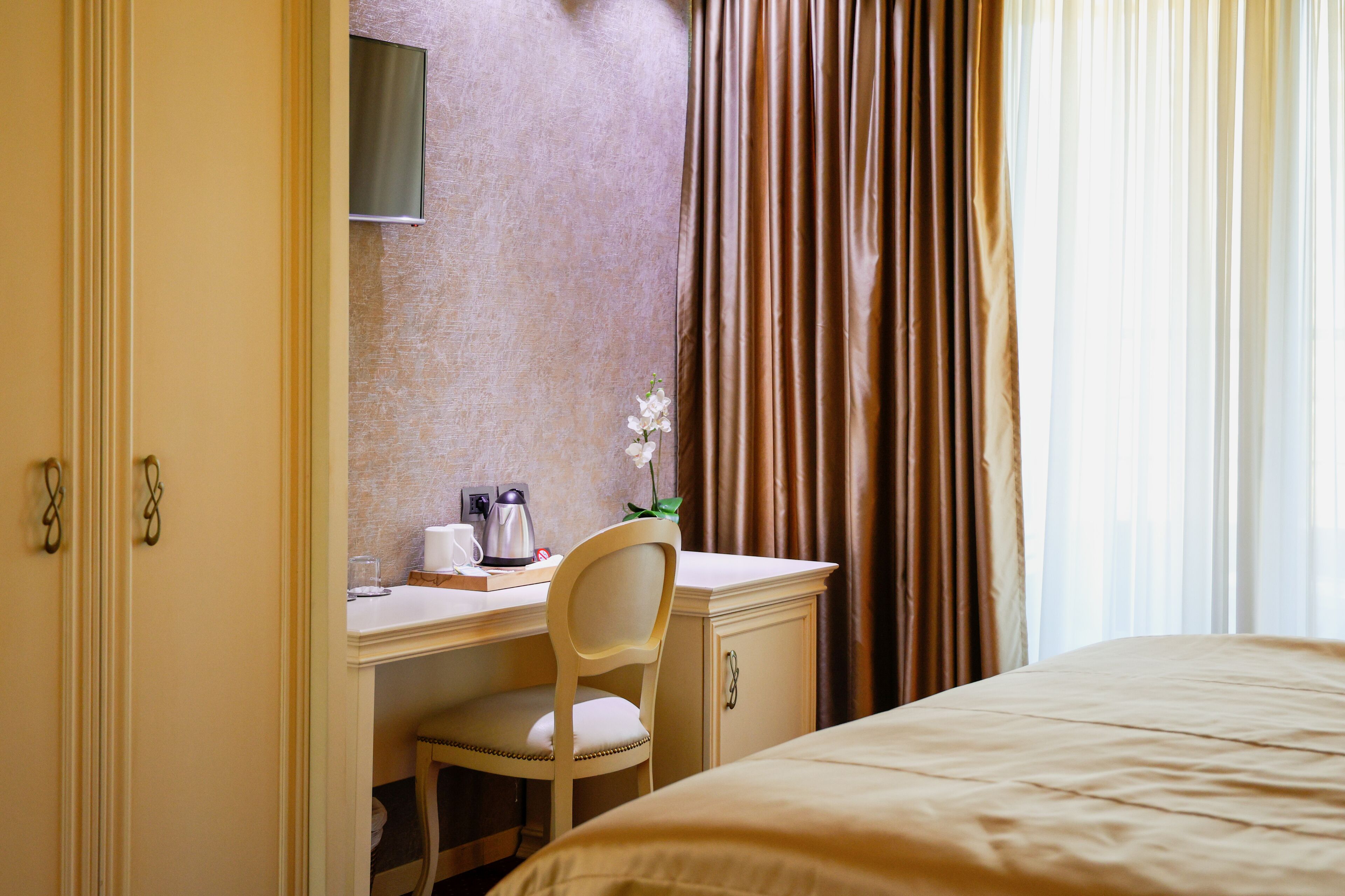 A hotel room with a bed, a desk with a chair, a small table with a coffee maker, and a window with curtains.