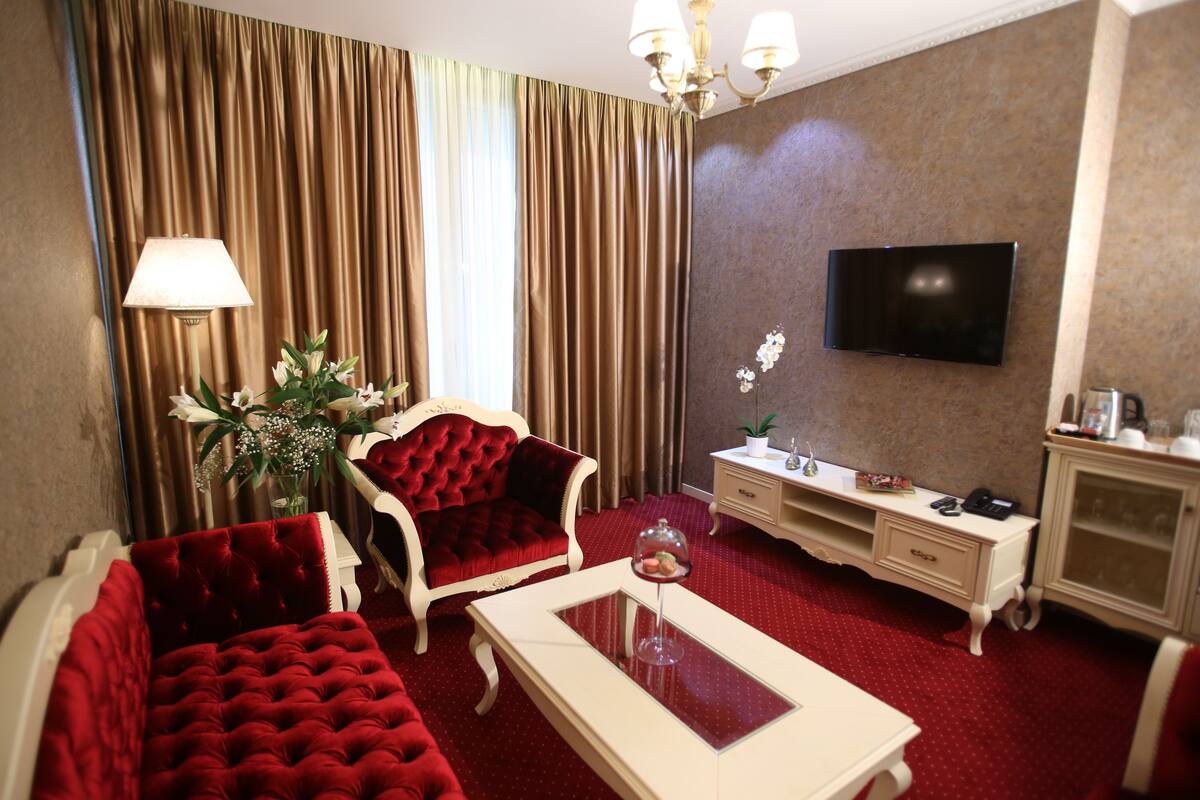 A hotel room with a red carpet, a TV mounted on the wall, a desk with a lamp, and a sofa.