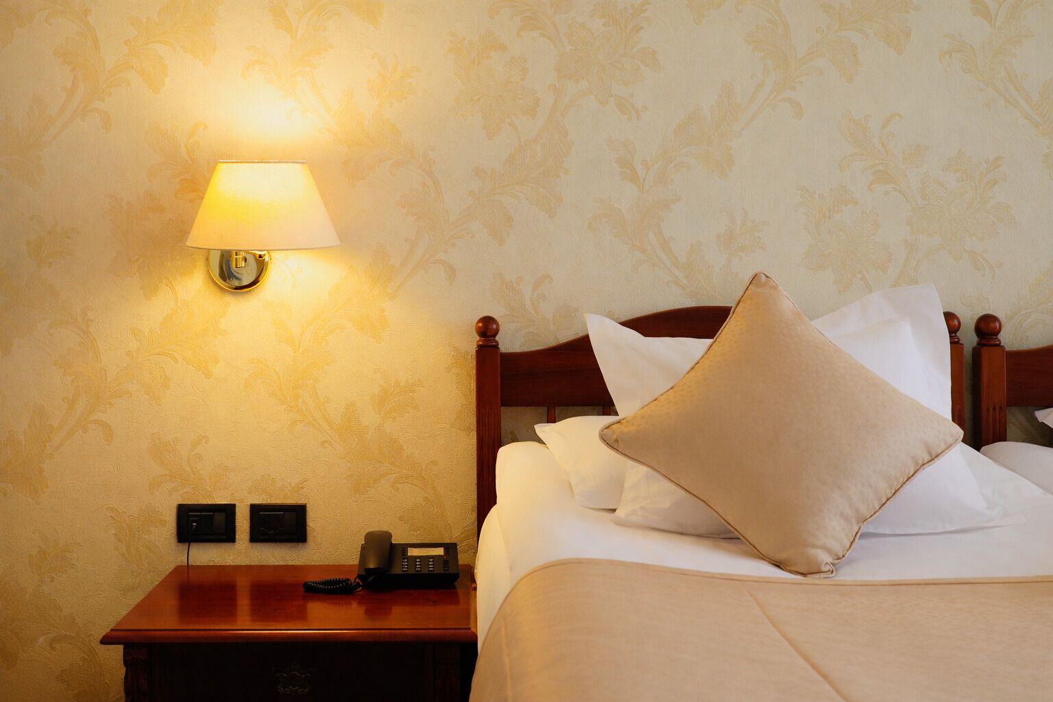 A hotel room with a wooden nightstand, a lamp, a telephone, and a bed with white bedding and a beige pillow.
