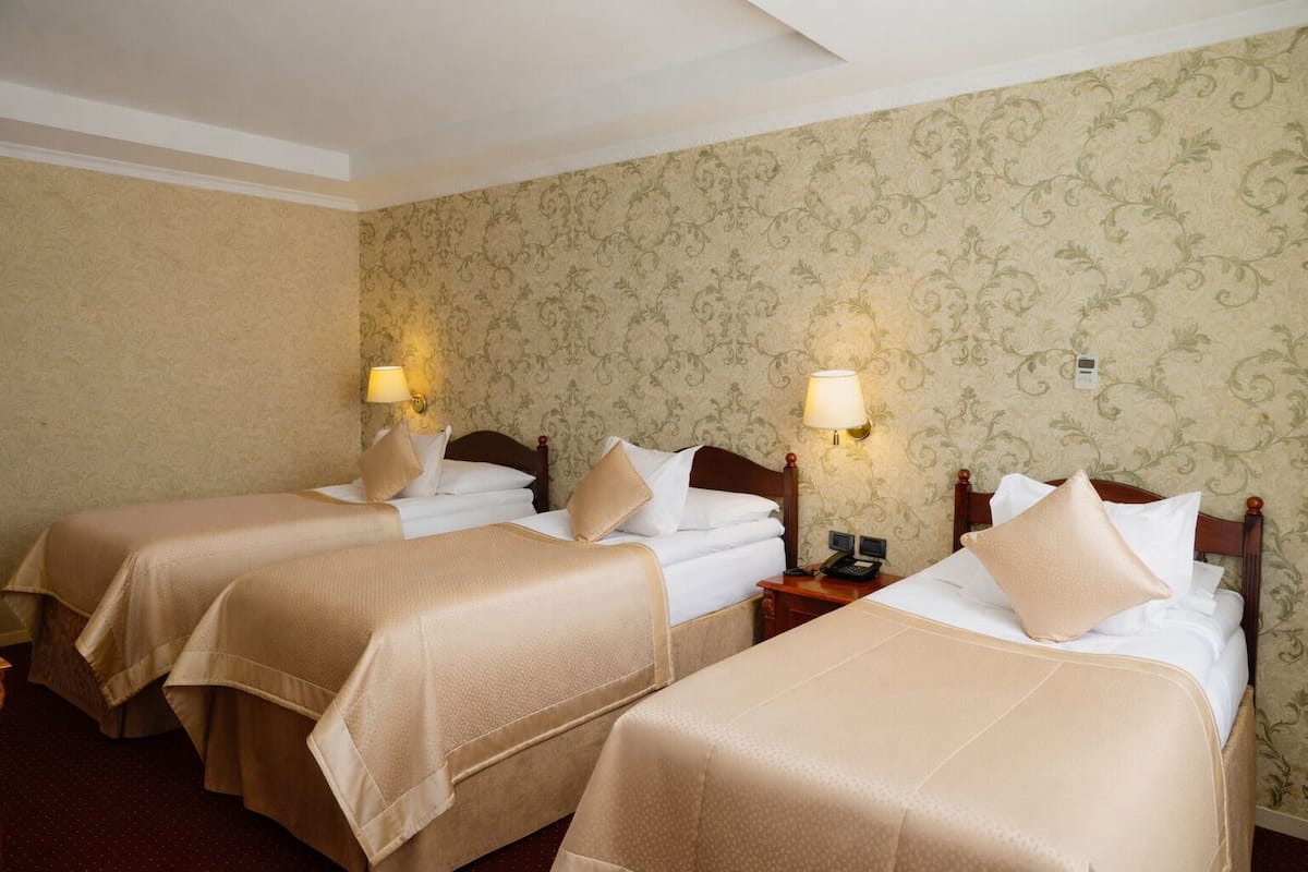 A hotel room with three beds, each with a wooden headboard and beige bedding. There are wall-mounted lamps on either side of the beds.