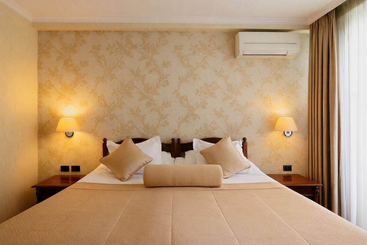 A hotel room with a large bed, two bedside lamps, an air conditioning unit, and patterned wallpaper.