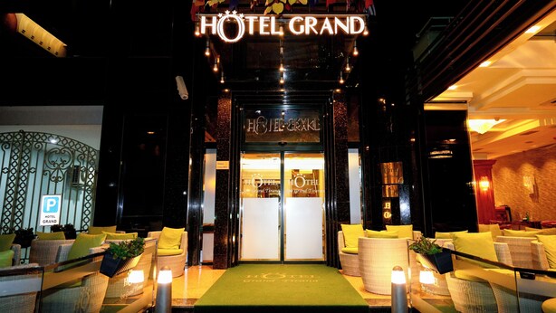 The entrance of HOTEL GRAND with a green carpet, wicker furniture, and a sign above the doors.