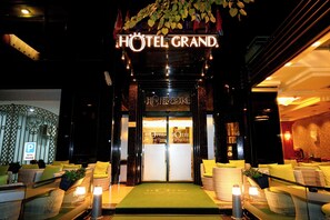 La entrada de HOTEL GRAND, con una alfombra verde, muebles de mimbre y un letrero sobre las puertas.