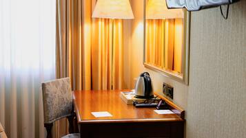A hotel room with a wooden desk, a chair, a lamp, a kettle, and a mirror.