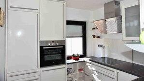 A modern kitchen with white cabinetry, a built-in oven, a window with blinds, and a stainless steel range hood. - Holiday house Karin Tassius - Holiday Karin Tassius (Vöhl)