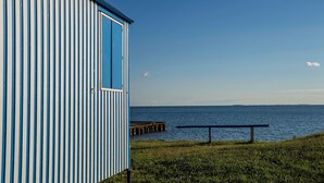 A coastal building with a blue and white striped exterior, a grassy area, a pier extending into the water, and a clear sky. - Penthouse sea view - Apartment sea view in the penthouse (Göhren)