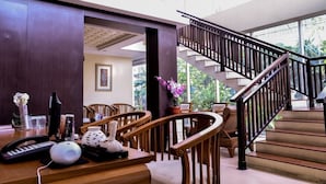 A spacious room with a wooden desk, chairs, a telephone, and a potted plant. A staircase with a railing leads to an upper level. - Grand Allison (Sentani)