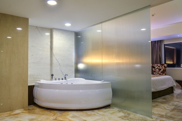 A modern bathroom with a large, round jacuzzi tub, a glass partition, and a bed with a patterned headboard in the adjacent room. - Grand Allison (Sentani)