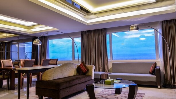 A modern hotel room with a large window, a sofa, a dining table, and a coffee table. - Grand Allison (Sentani)