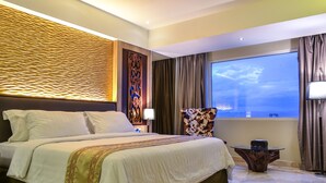 A hotel room with a large bed, a nightstand, a table, and a view of the sky through the window. - Grand Allison (Sentani)