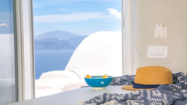 Una habitación con vistas al mar y a las montañas, un cuenco azul con limones, un sombrero de paja y una manta estampada sobre la cama.
