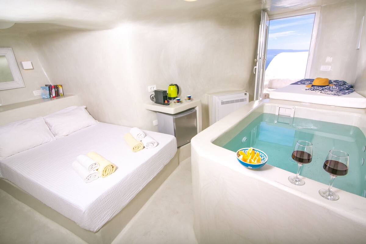 A room with a bed, a small table with a kettle, a window with a view, and a bathtub with glasses and a bowl.