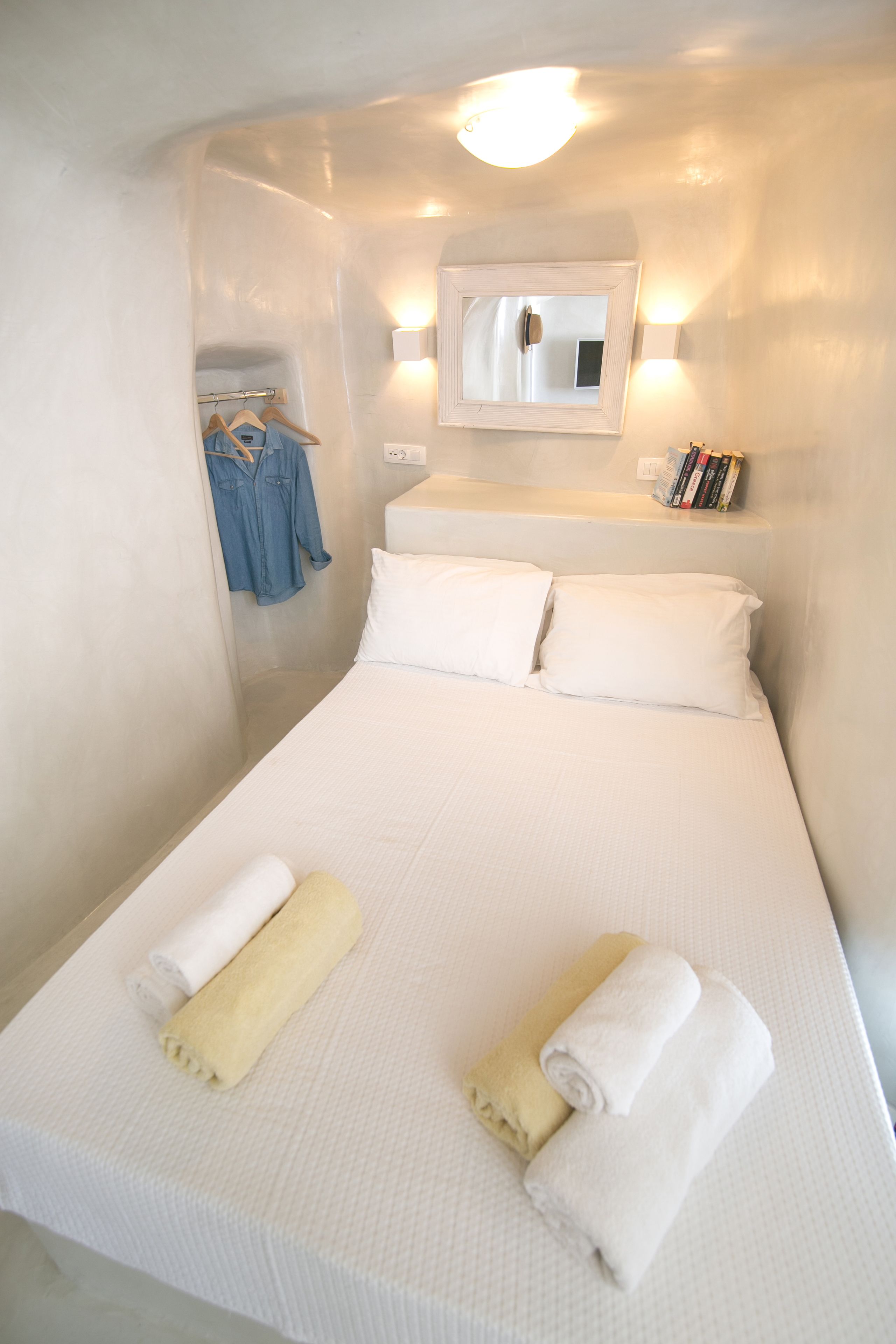 A small, neatly made bed with white linens and two rolled towels, a wall-mounted mirror, and a shelf with books and a small clock.