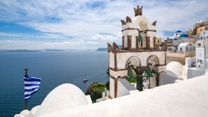 Vista costera con un edificio blanco destacado que cuenta con un campanario, sobre el mar con barcos y cielo despejado.