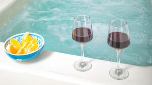 Una habitación con una bañera, un cuenco de naranjas y dos copas de vino tinto.