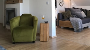 Een woonkamer met een groene fauteuil, een houten bijzettafeltje met een glas en een bank met een plaid.
