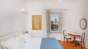 A bedroom with a bed, a window with a view of a building, a table with chairs, and a wall-mounted air conditioner. - Apartment Entre Les Jardins (Dubrovnik)